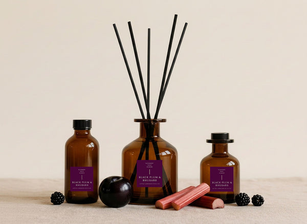 Plum & Rhubarb reed diffusers in three sizes, amber glass with deep purple labels and black reeds, styled with blackberries, a dark plum and cut rhubarb stems