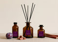 Load image into Gallery viewer, Plum & Rhubarb reed diffusers in three sizes, amber glass with deep purple labels and black reeds, styled with a fresh plum and rhubarb stems
