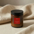 Load image into Gallery viewer, Christmas Spice candle in amber glass with its black lid on, resting on a cream knitted throw