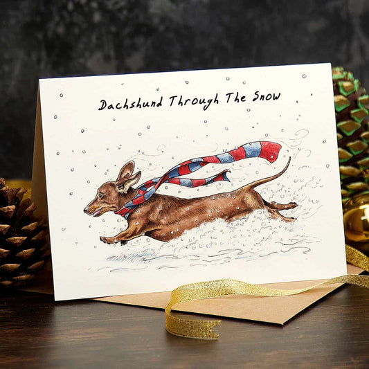 Christmas card titled 'Dachshund Through The Snow', with a dachshund in a striped scarf running through snow