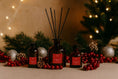 Load image into Gallery viewer, Christmas Spice reed diffusers among evergreen foliage, baubles and berries with fairy lights behind
