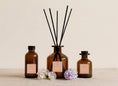 Load image into Gallery viewer, Bouquet reed diffusers in three sizes, amber glass with blush pink labels and black reeds, styled with sweet pea and lilac blooms
