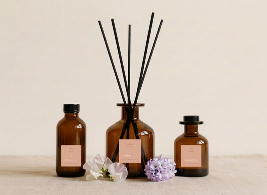 Bouquet reed diffusers in three sizes, amber glass with blush pink labels and black reeds, styled with sweet pea and lilac blooms