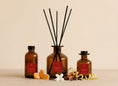 Load image into Gallery viewer, Christmas Spice reed diffusers in three sizes, amber glass with deep red labels and black reeds, styled with satsuma segments, cinnamon sticks, cloves and citrus peel
