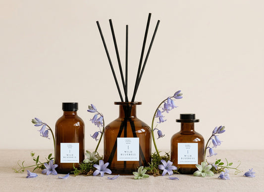 English Bluebell reed diffusers in three sizes, amber glass with pale blue labels and black reeds, styled with fresh bluebells and moss