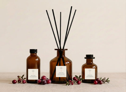 Festive Cranberry reed diffusers in three sizes, amber glass with cream labels and black reeds, styled with frosted cranberries and winter foliage