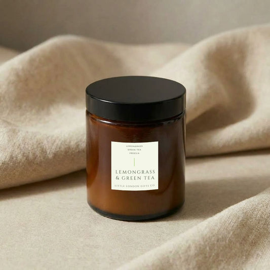 Lemongrass & Green Tea candle in amber glass with its black lid on, resting on cream fabric