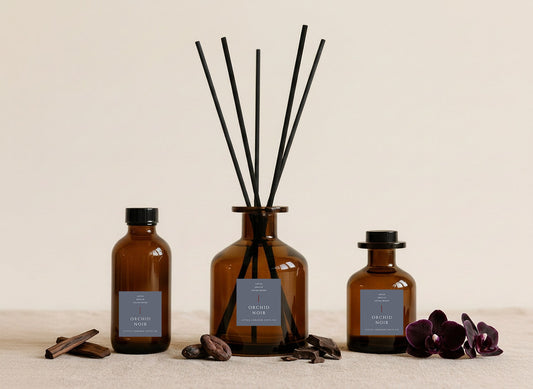 Orchid Noir reed diffusers in three sizes, amber glass with slate grey labels and black reeds, styled with cocoa beans, sandalwood and dark orchids