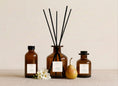 Load image into Gallery viewer, Pear & Freesia reed diffusers in three sizes, amber glass with cream labels and black reeds, styled with a ripe pear and white freesia blooms
