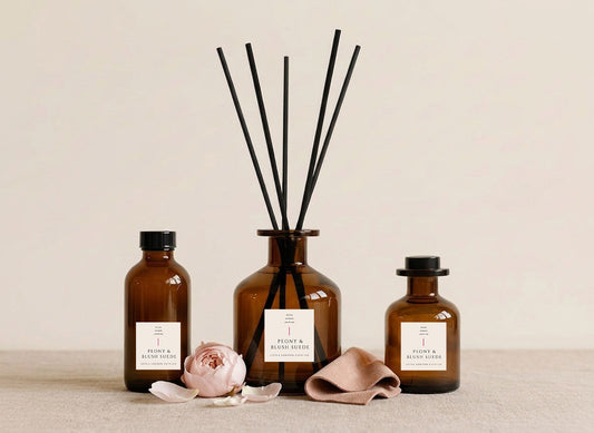 Peony & Suede reed diffusers in three sizes, amber glass with cream labels and black reeds, styled with a blush peony and folded suede