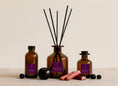 Load image into Gallery viewer, Plum & Rhubarb reed diffusers in three sizes, amber glass with deep purple labels and black reeds, styled with blackberries, a dark plum and cut rhubarb stems
