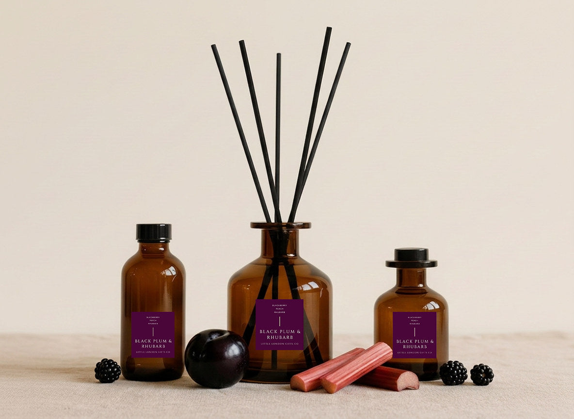 Plum & Rhubarb reed diffusers in three sizes, amber glass with deep purple labels and black reeds, styled with blackberries, a dark plum and cut rhubarb stems