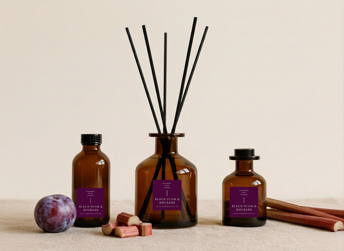 Plum & Rhubarb reed diffusers in three sizes, amber glass with deep purple labels and black reeds, styled with a fresh plum and rhubarb stems