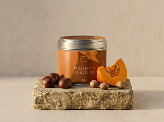 Pumpkin & Chestnut candle tin in brushed silver with an amber label, styled with chestnuts, nutmeg and a wedge of pumpkin on stone