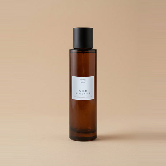 English Bluebell room spray 100ml, amber glass bottle with a matte black cap and pale blue label, on a warm beige backdrop