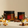 Load image into Gallery viewer, Christmas Spice candles in two sizes, amber glass with black lids and deep red labels, styled with dried orange, cinnamon and pine cones
